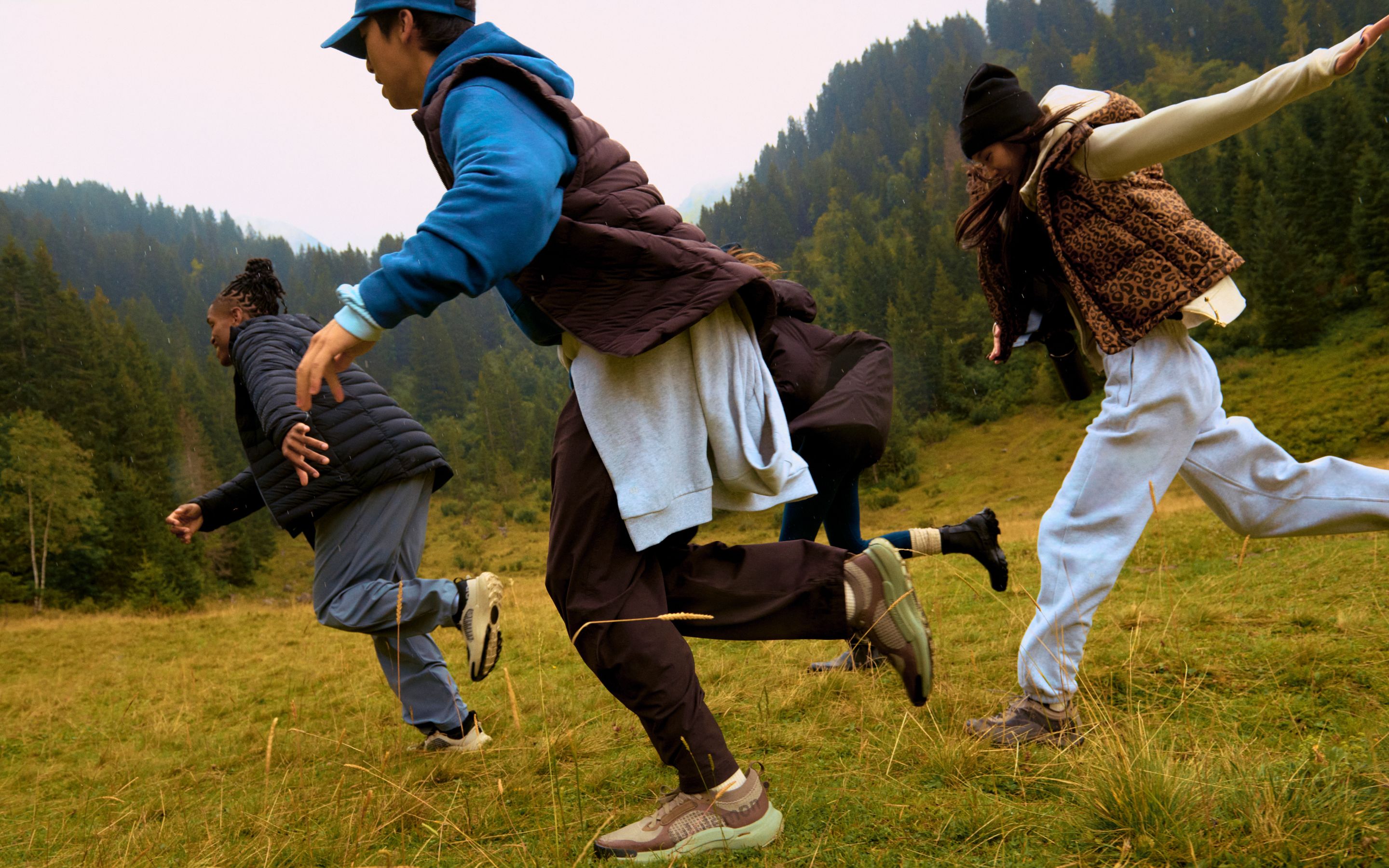 Three models running in front of forested mountains: one wearing the Navigation Down Hoodie in black, one in the Navigation Down Vest, and one in the Wunder Puff Vest in leopard print. 
