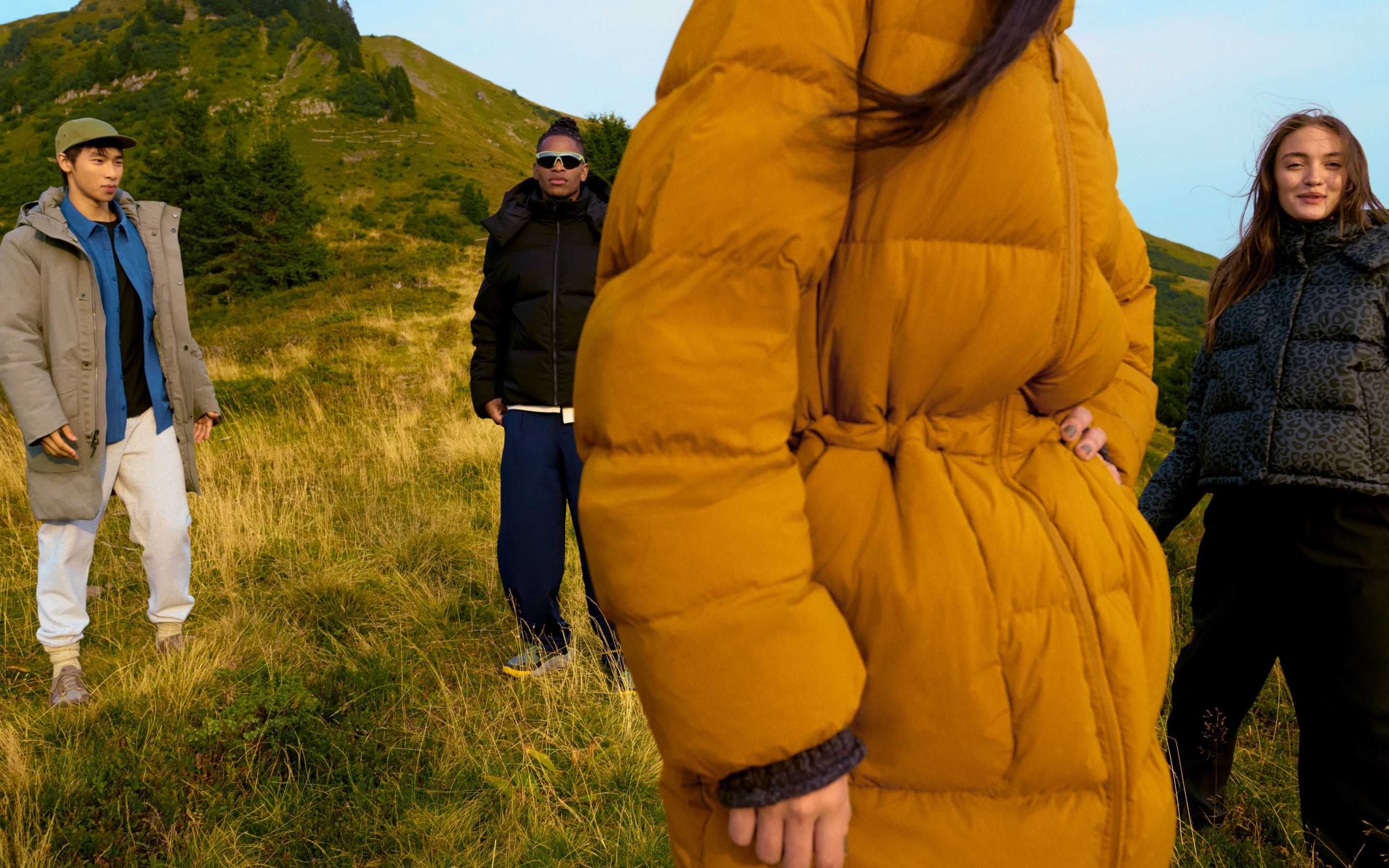 Models in lululemon outerwear standing on a grassy hillside: one in the grey Waterproof Parka, one in the black Navigation Down Hoodie, one in the tan Insulated Cinch Puff Jacket, and one in the grey leopard print Wunder Puff Jacket. 