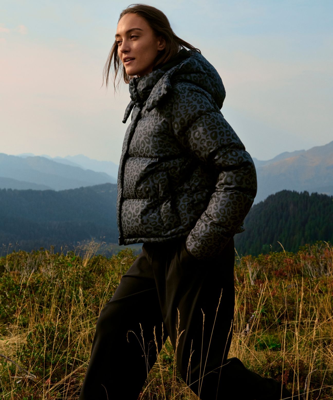 Model outdoors on a grassy hillside wearing the grey leopard print Wunder Puff Jacket with hands in pockets, mountains in the distance.