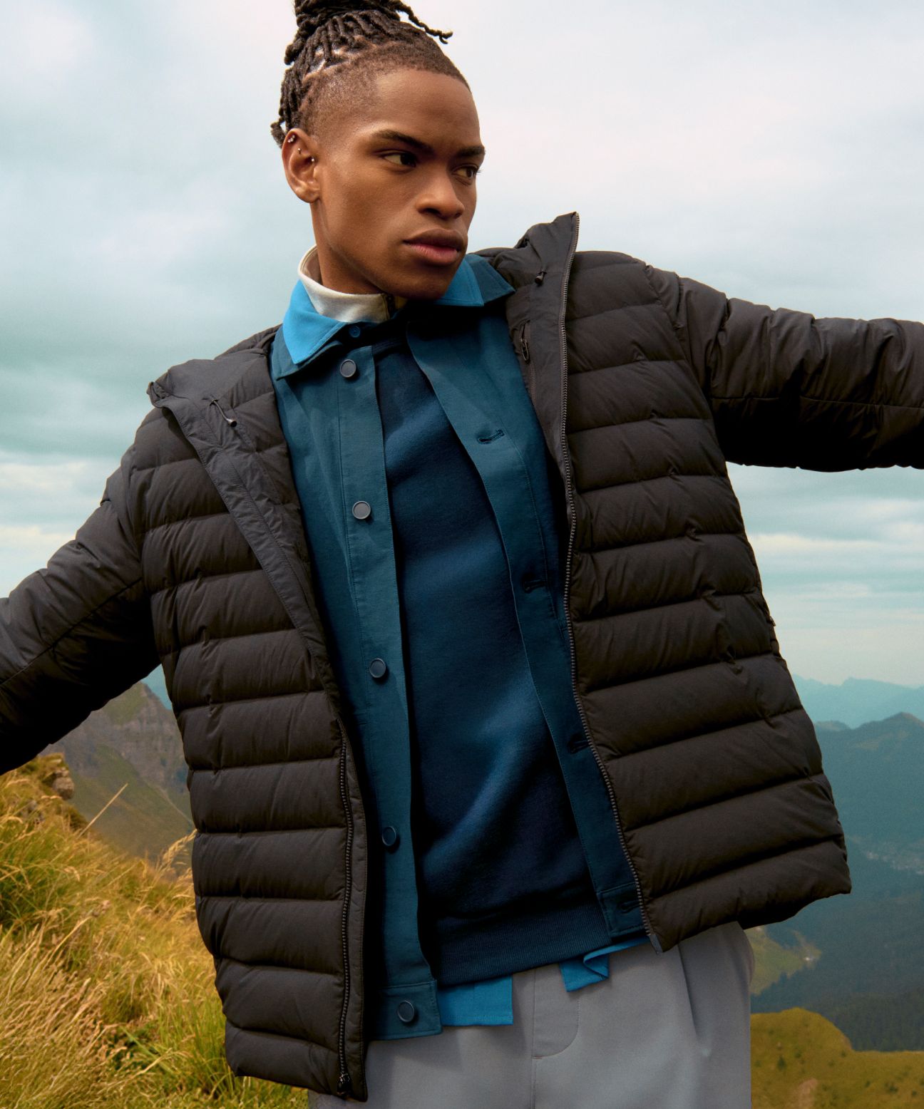 Model outdoors wearing the black Navigation Down Hoodie layered over a blue shirt, standing in front of rural greenery.