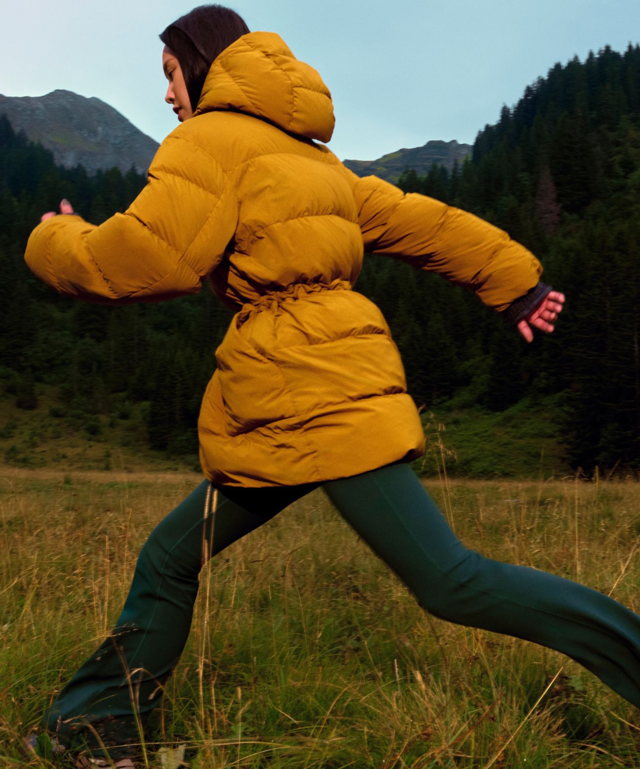 Model running across grass in the Insulated Cinch Puff Jacket in tan, mountains visible in the background.
