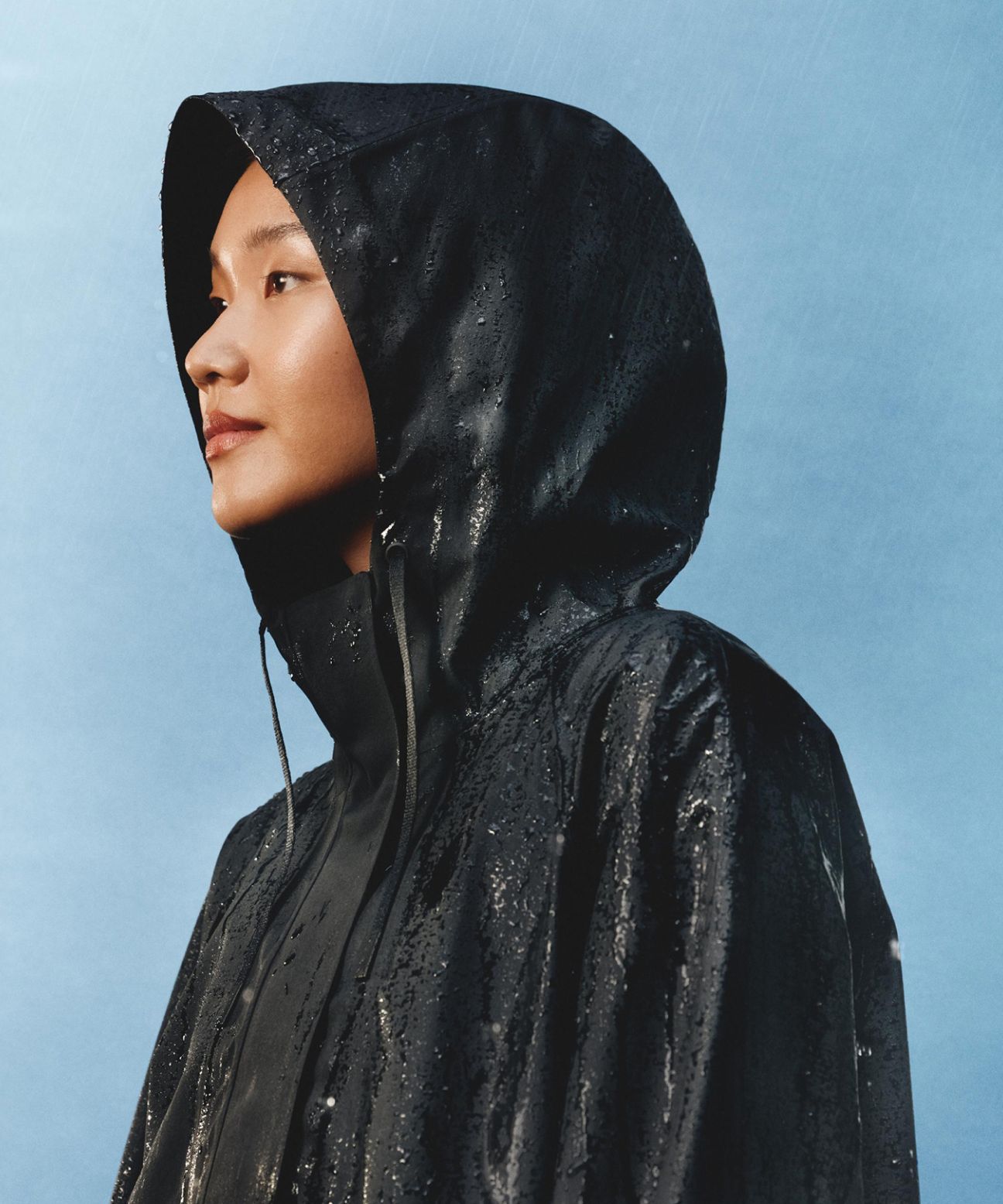A person wearing the Fishtail Rain Coat with the hood up staring to the side while rain falls around them.