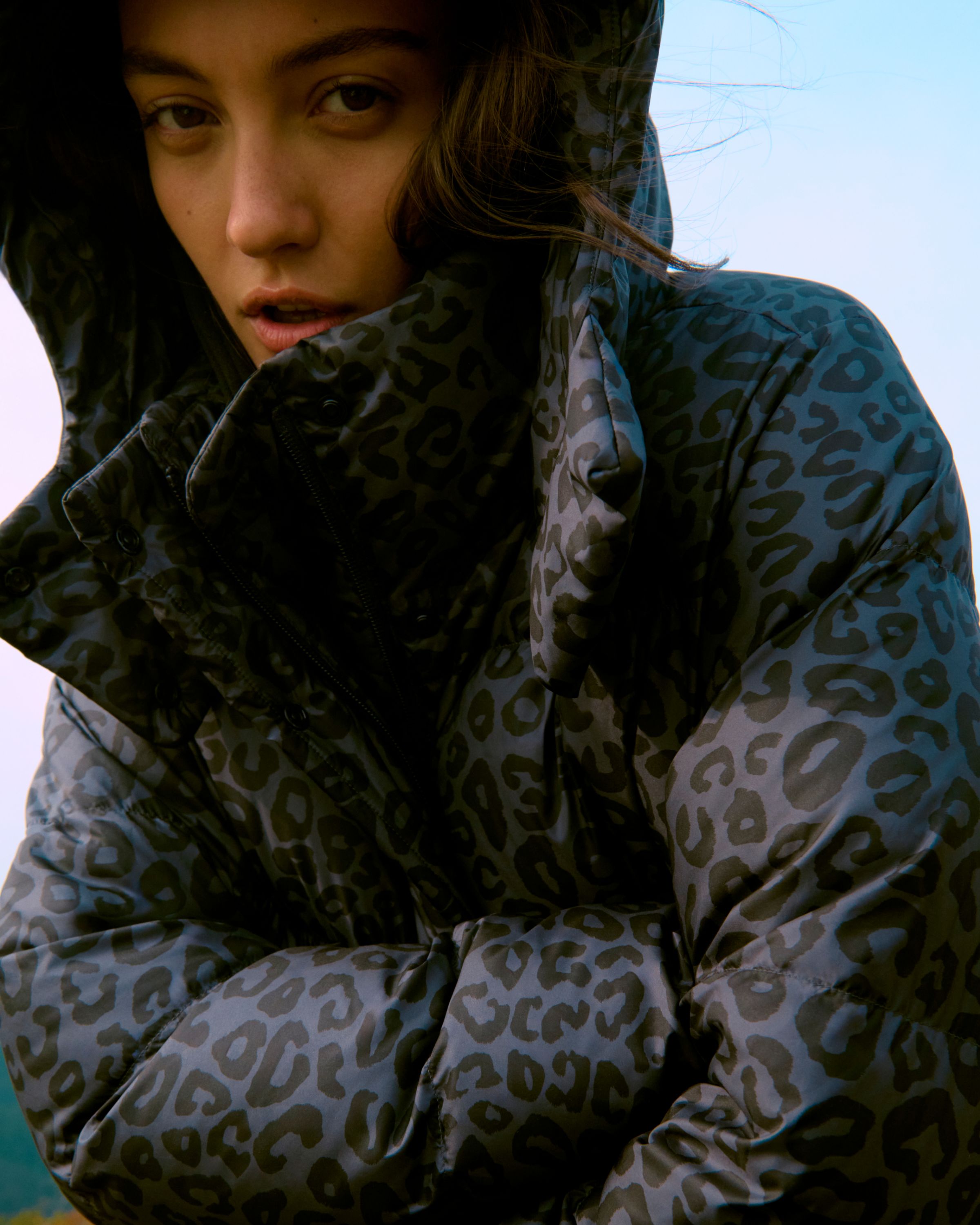 Close-up of a model wearing the Wunder Puff Jacket in grey leopard print with the hood up against a pale sky backdrop.