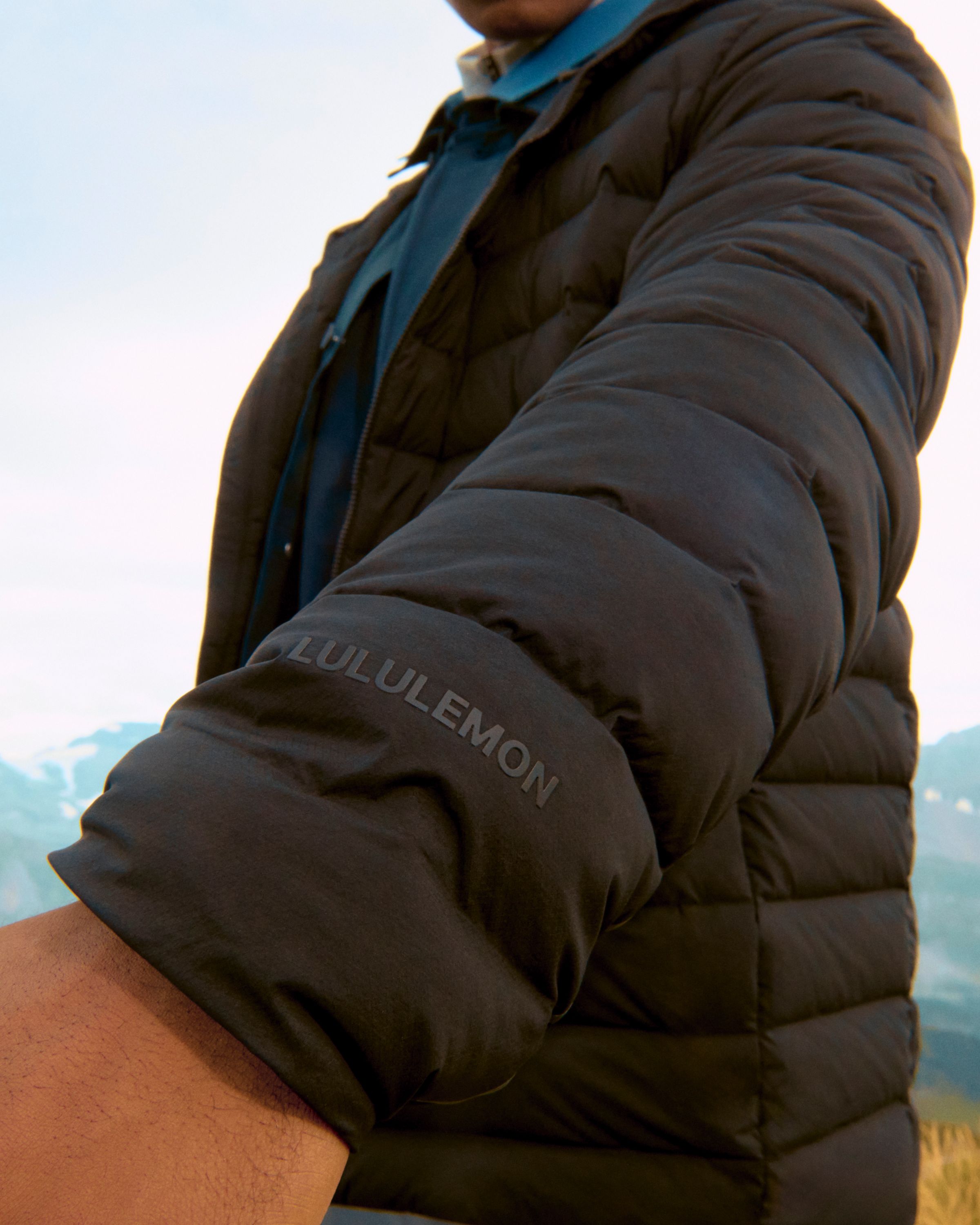 Close-up of a black Navigation Down Hoodie sleeve with the lululemon logo visible and mountains blurred in the background.