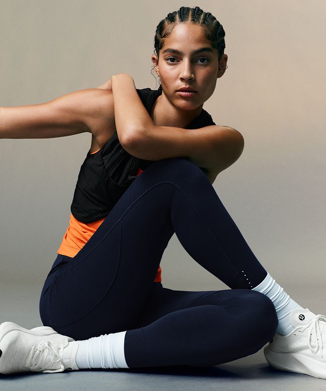 A person wearing a Fast and Free Running Top in Orange Punch, a Fast and Free Running Vest in Black, and the Fast and Free High-Rise Tight 25” in True Navy.