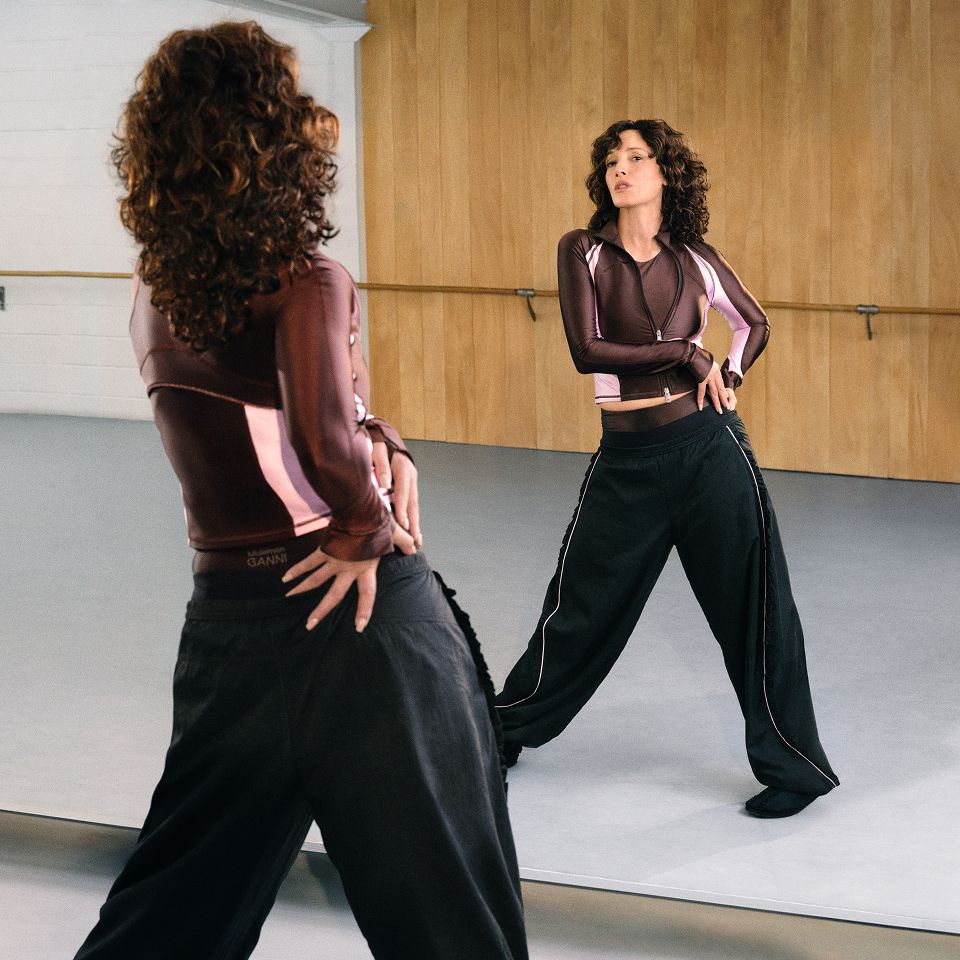 A person posing in front of a dance studio mirror wearing the Define Ruffle Trim Jacket and the Trim Textured Pant, from the GANNI x lululemon collection.