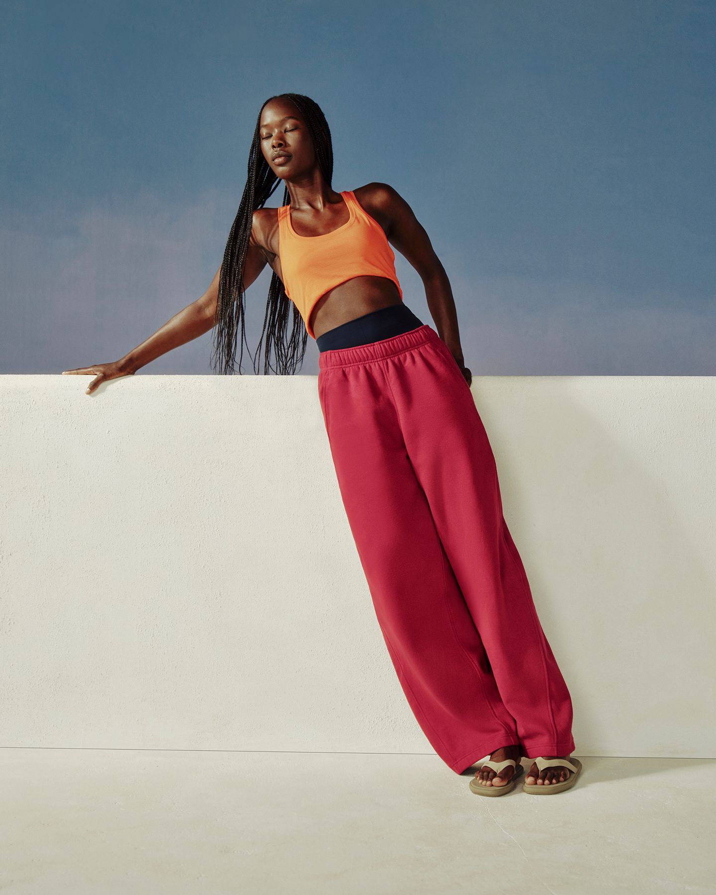 A person leans again a white ledge wearing the new Scuba Evolve Straight Leg Pant in Raspberry Rave and Swiftly Tech Tank 2.0 in Orange Crush.
