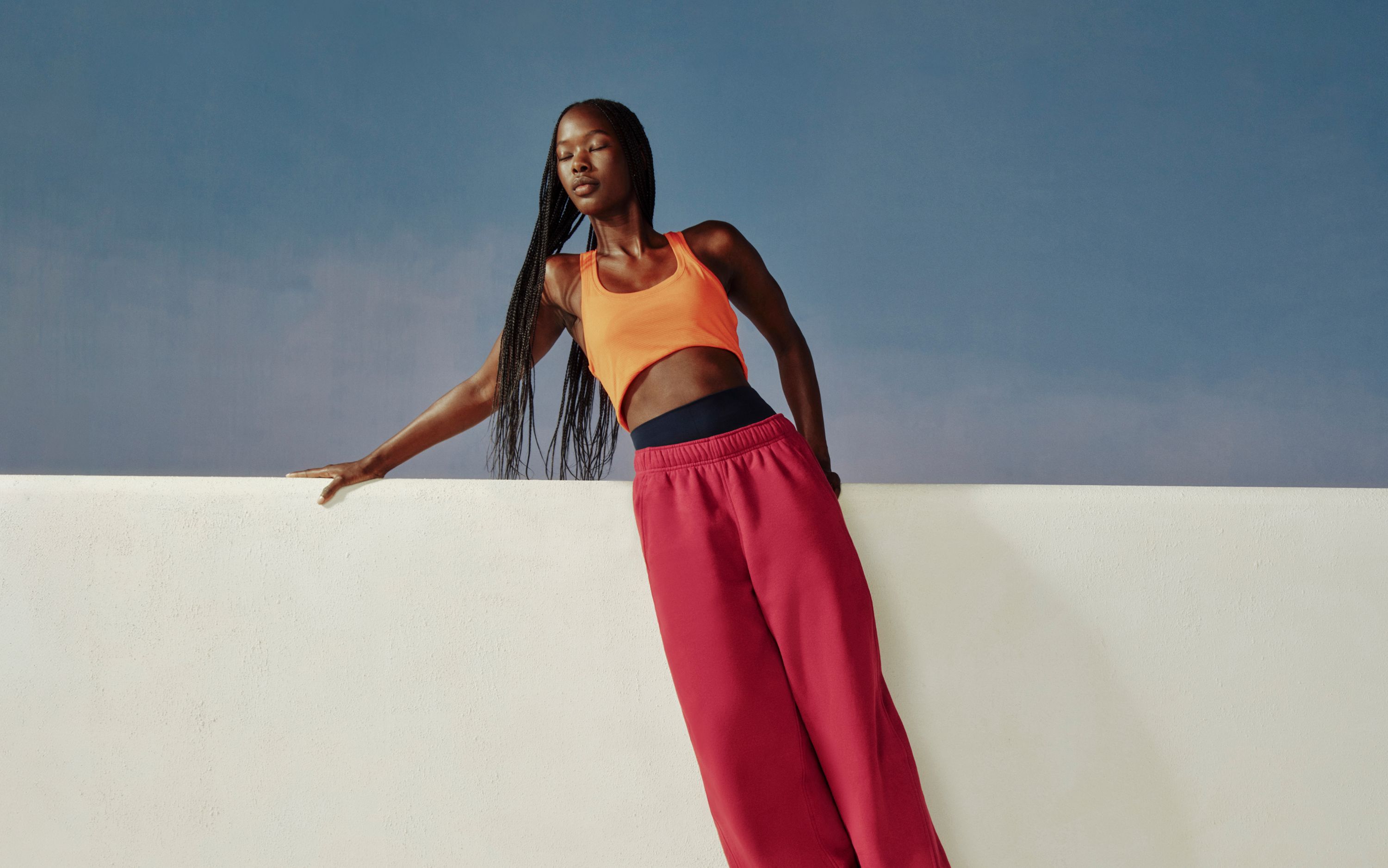 A person leans again a white ledge wearing the new Scuba Evolve Straight Leg Pant in Raspberry Rave and Swiftly Tech Tank 2.0 in Orange Crush.