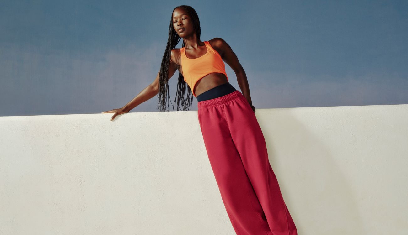 A person leans again a white ledge wearing the new Scuba Evolve Straight Leg Pant in Raspberry Rave and Swiftly Tech Tank 2.0 in Orange Crush.