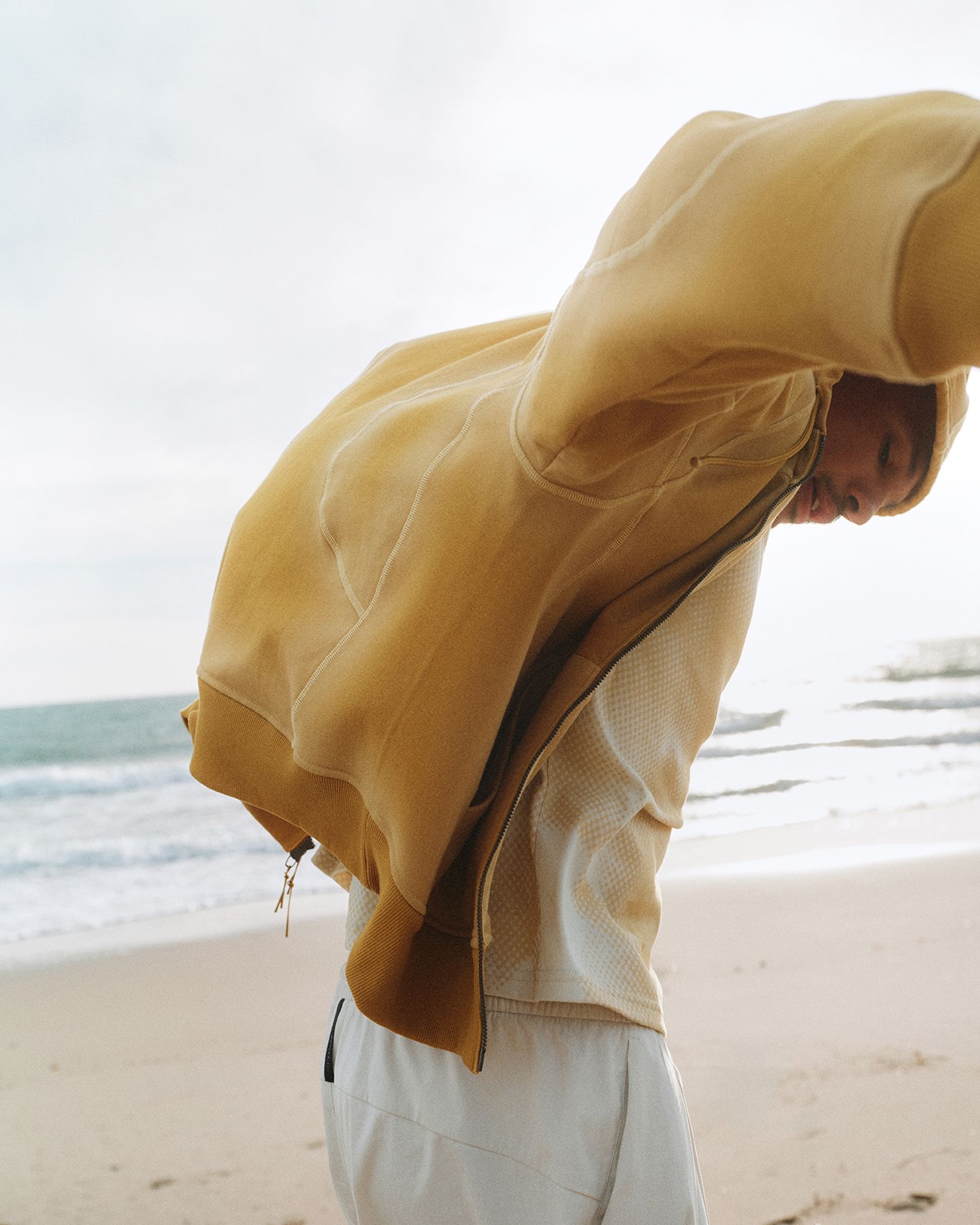A person walks along the beach with their head down. They are wearing a sweater from the summer 2026 SLSNH collection that is blowing in the wind behind them.