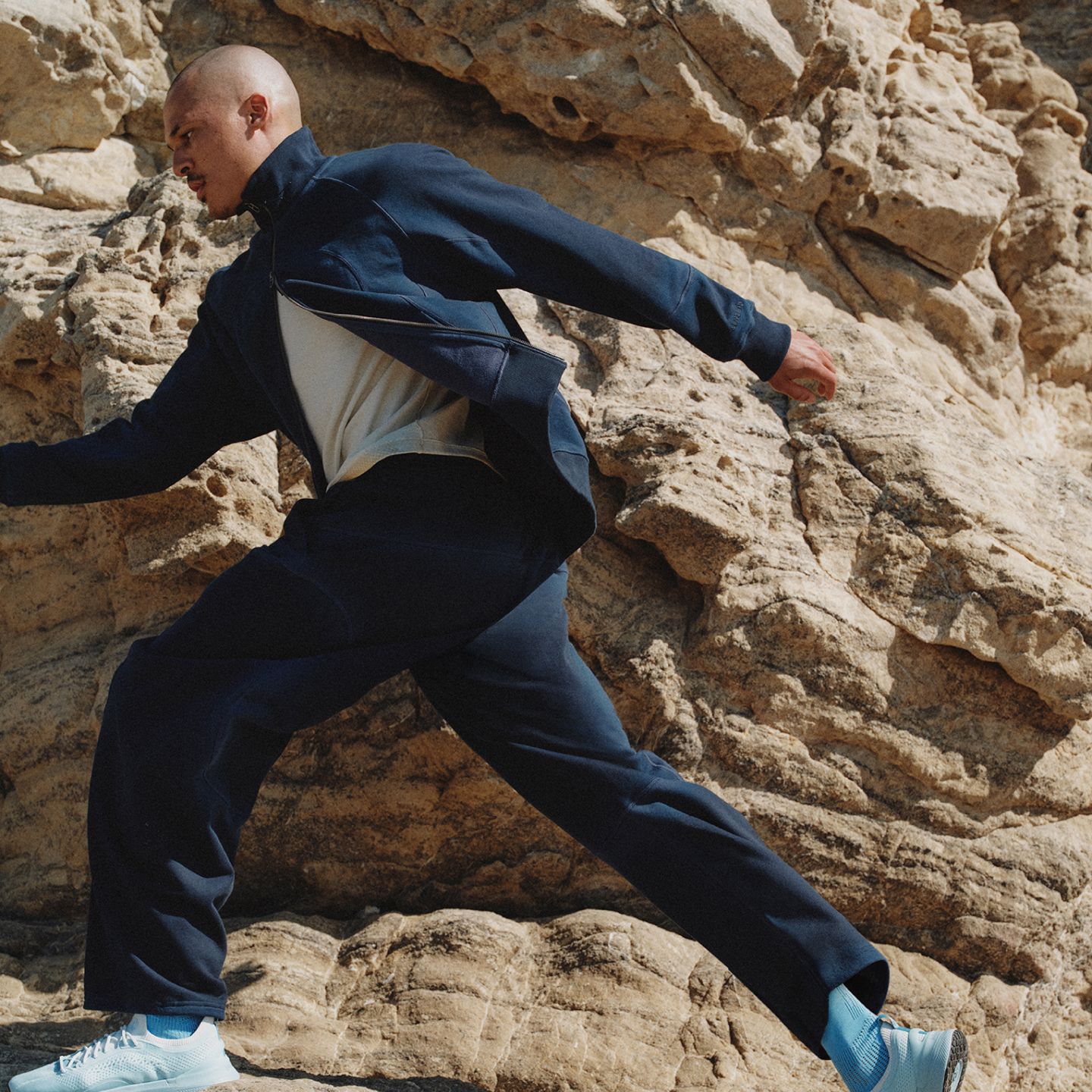 A person in motion climbs a along a large rock wearing clothes from the summer 2026 SLNSH collection.