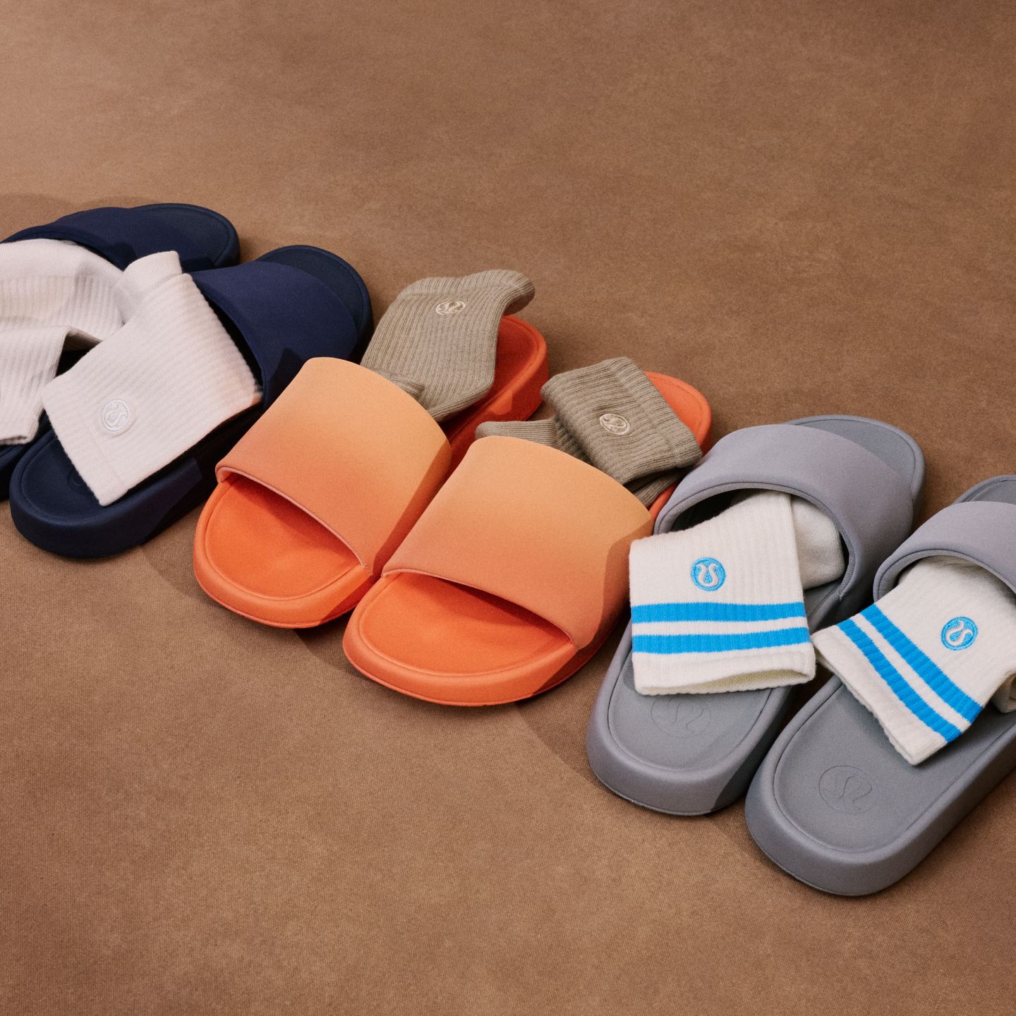 Trois paires de sandales Restfeel alignées en différentes couleurs, avec une paire de bas glissée dans chaque paire, devant un fond mauve.