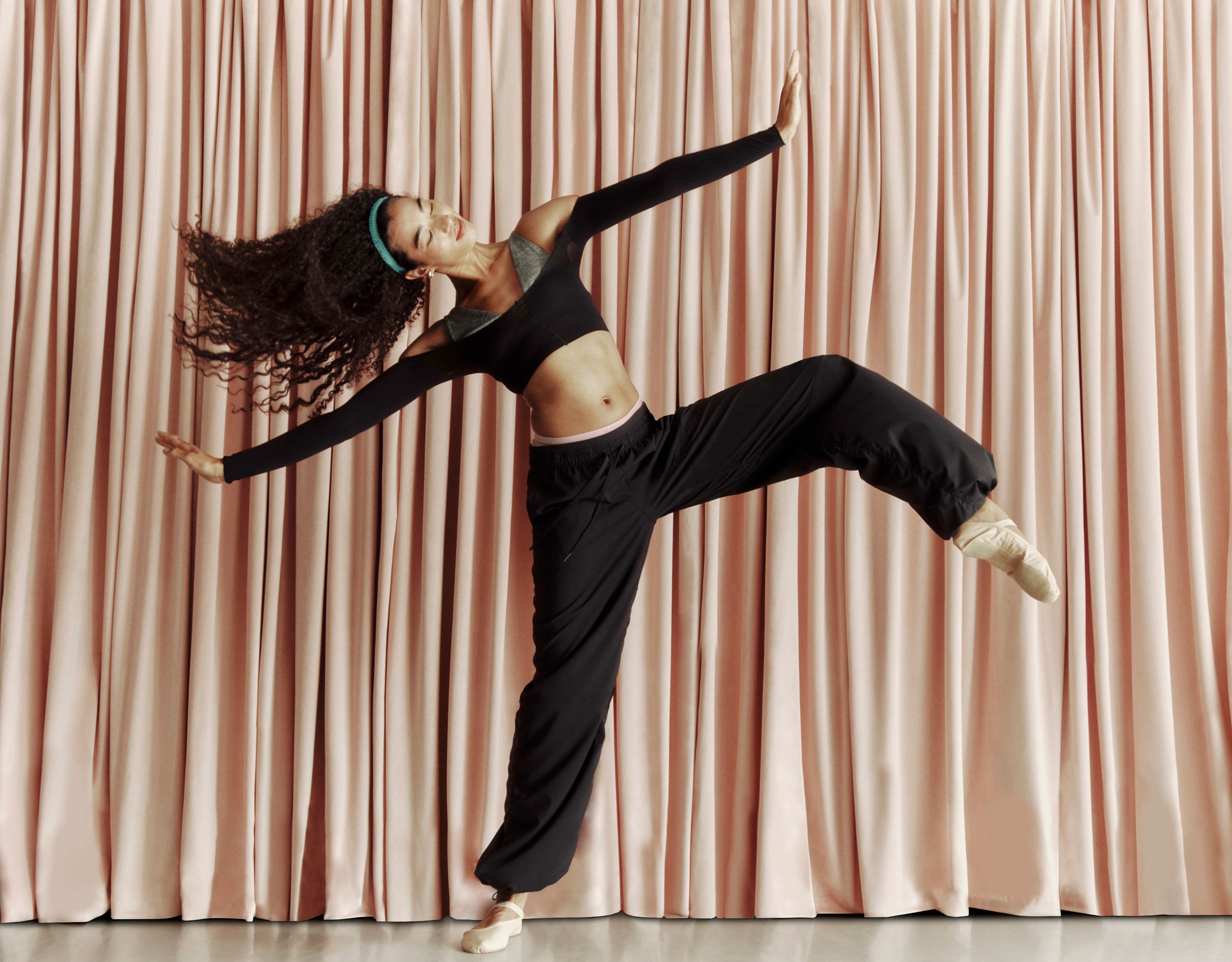 A person is mid-movement with their arms outstretched in front of a pink curtain. They are wearing the Dance Studio Evolve Low-Rise Oversized Pant in and the Nulu Triangle Bralette A-C.