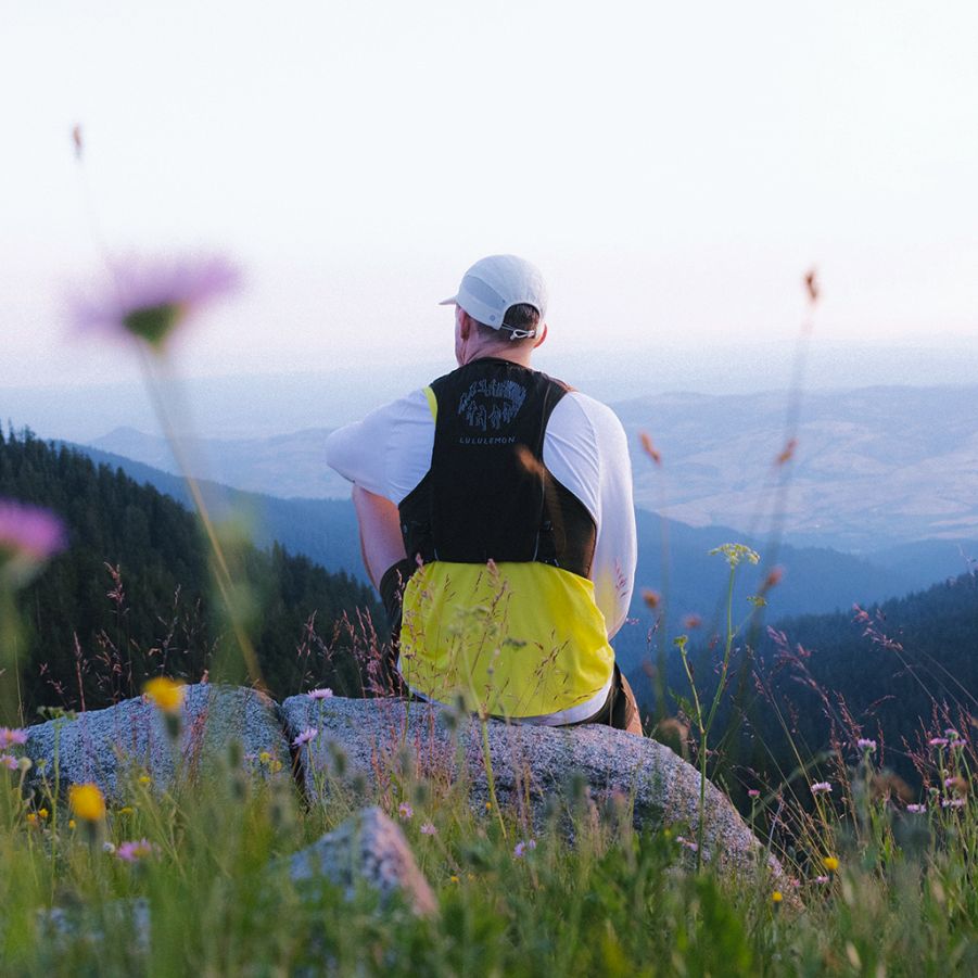 A person in a lululemon x Geoff McFetridge shirt and vest with 'Amplify the Quiet' graphic, sits on a rock overlooking mountains, pine forests, and valleys.