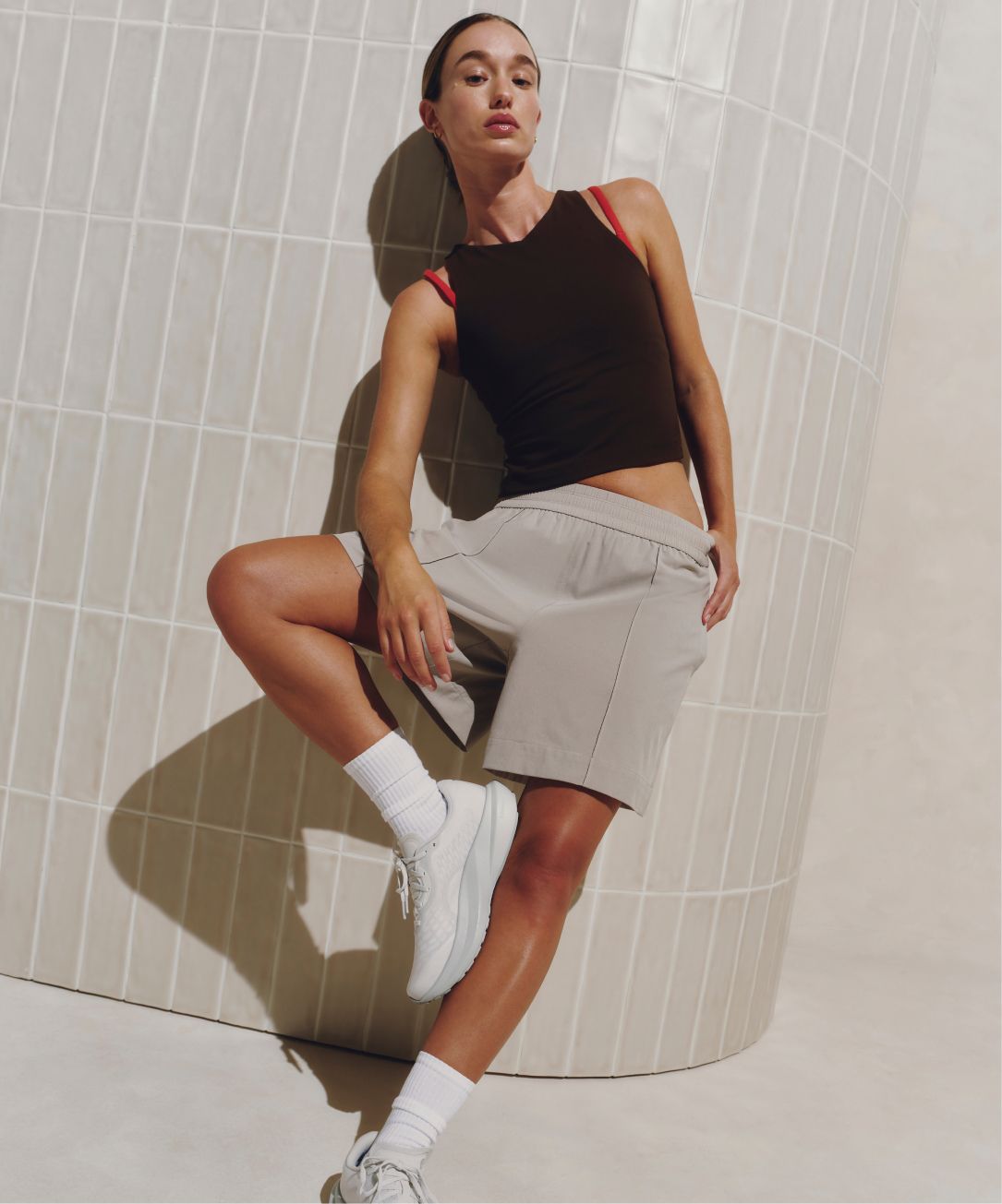 A person poses in front of a tiled wall with their one leg propped up against the other. They are wearing the Breezily Shorts in Downtown Tan and Align Mini V-Neck Tank in Walnut Crunch. 