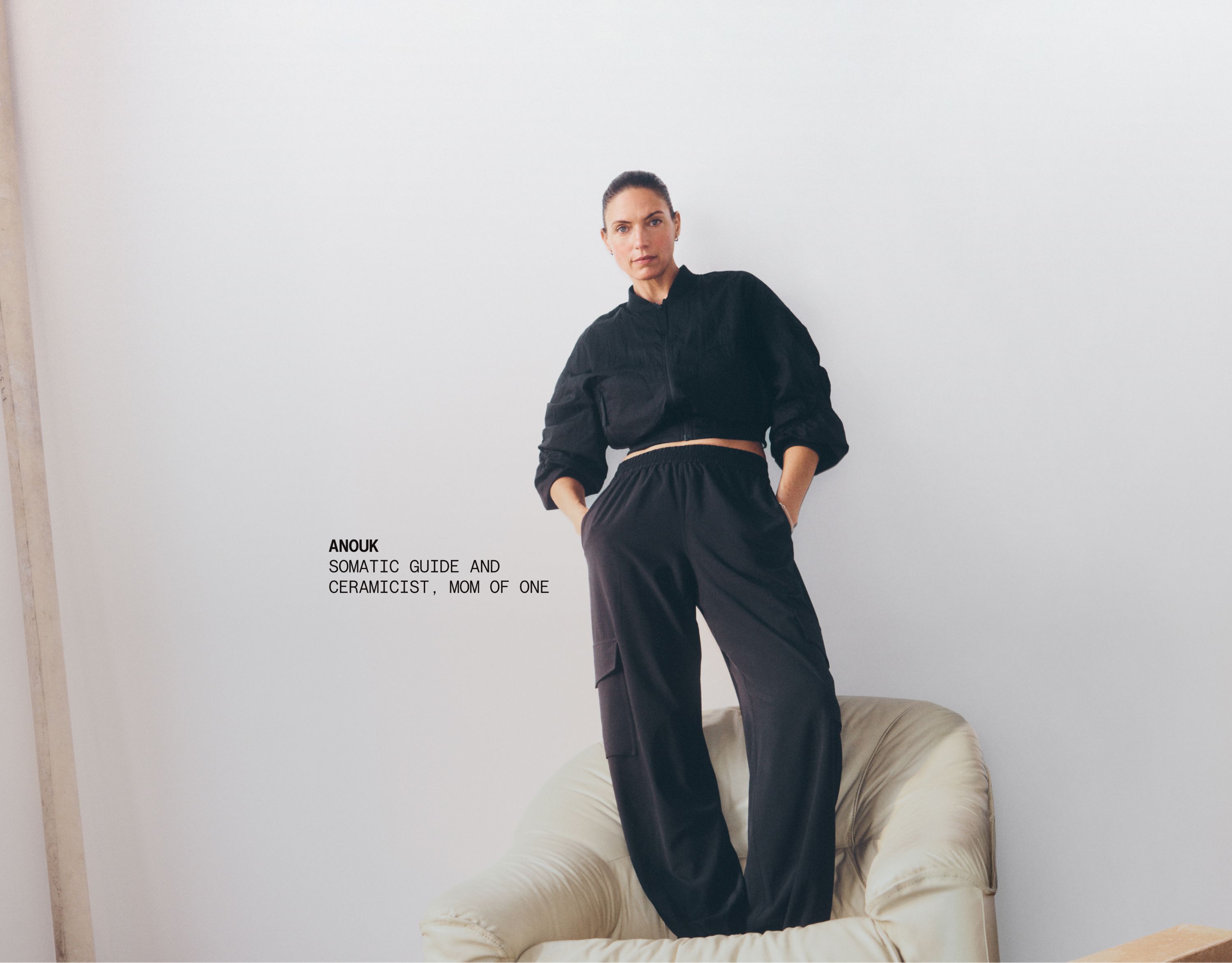 A person wearing the lululemon Curved Hem Cropped Bomber and Breezily Cargo Pant stands on a beige chair against a neutral backdrop. Text overlay reads: Anouk, Somatic guide and Ceramicist, Mom of one. 