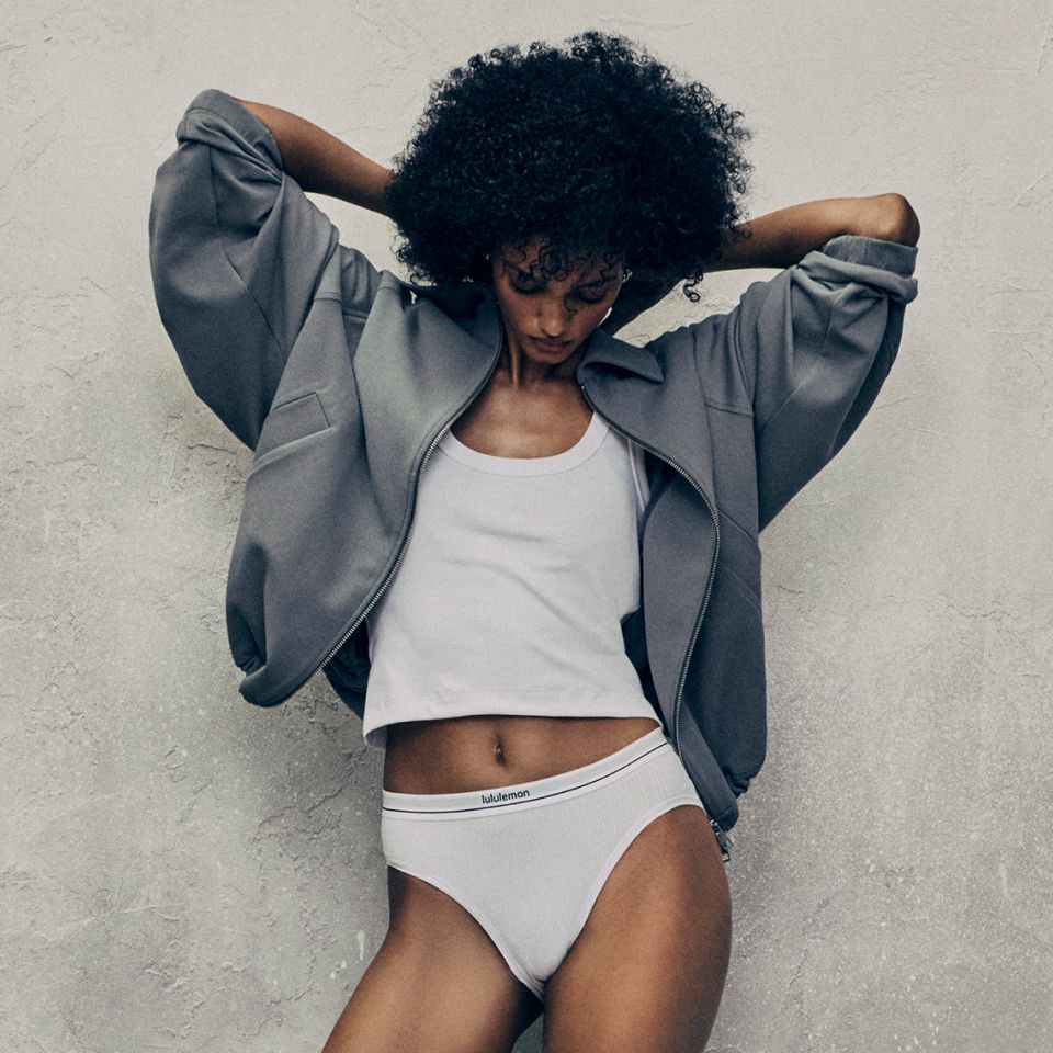  A person poses against a grey wall. They are looking down and have their hands behind their head slightly adjusting the back of their collar. They are wearing new styles from the fall 2026 line. 