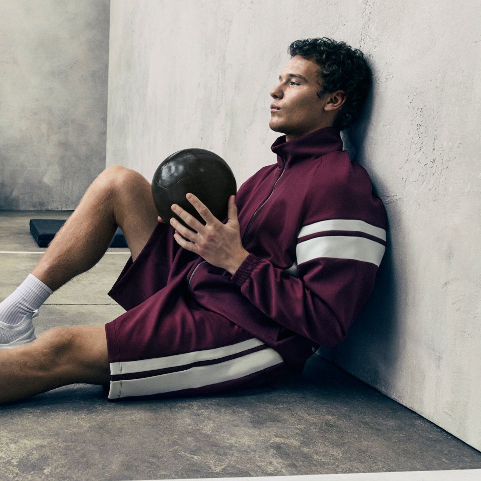  A person sits leaning against a grey wall in a studio. They have one leg bent towards them and the other outstretched. They are holding a brown ball and wearing new styles from the fall 2026 line.